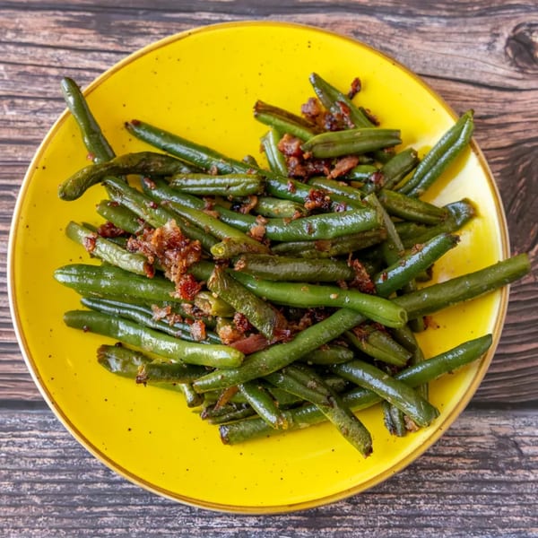 Green beans with bacon bits on a bright yellow plate, on a rustic wooden table.